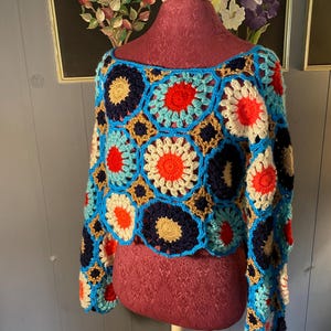 May include: A long-sleeved crochet top constructed from a series of interconnected circular and square motifs. The garment features a vibrant color palette including bright blue, deep navy, cream, orange, and red yarn in a repeating floral pattern.