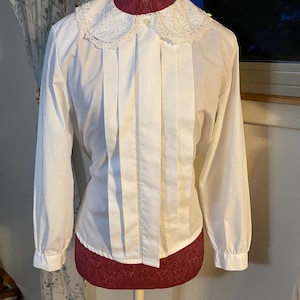 Puede incluir: Blusa blanca de manga larga con cuello decorativo con ribete de encaje. La blusa presenta pliegues verticales en la parte delantera y puños abotonados. La prenda se exhibe en un maniquí rojo.