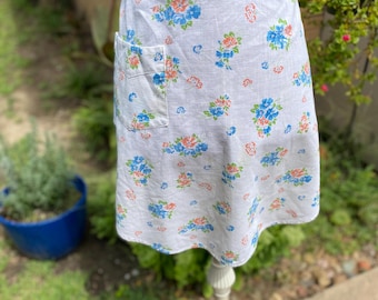 May include: White apron with a blue and orange floral pattern. The apron has a blue zigzag trim at the waist and a pocket on the front. The apron is displayed on a white stand in a garden setting.