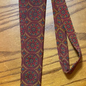 May include: A patterned necktie with a repeating floral design in red, teal, and gold. The tie is made of a soft fabric and is suitable for formal or casual wear. The tie is laying on a wooden surface.