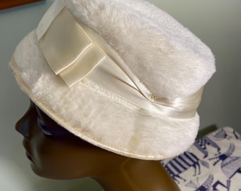 May include: A white faux fur pillbox hat featuring a smooth, cream-colored satin ribbon band wrapped around the base. The ribbon includes a decorative bow detail on the side. The hat has a flat top and a structured, rounded silhouette.