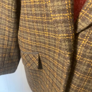 May include: A dark charcoal gray blazer featuring a prominent yellow grid pattern throughout the fabric. The garment includes a structured lapel, a single flap pocket on the side, and a deep red interior lining visible at the collar area.