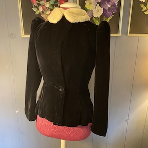 May include: A black velvet jacket featuring long sleeves with puffed shoulders, a single large black button closure at the waist, and a contrasting cream-colored faux fur collar. The jacket has a fitted silhouette and a slightly flared hemline.