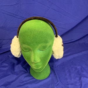 Puede incluir: Orejeras de piel sintética blanca con una diadema marrón. Las orejeras se llevan en una cabeza de maniquí verde.