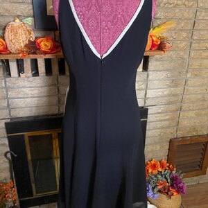 May include: A black sleeveless dress with a V-neckline and white trim. The dress is displayed on a pink dress form. The dress has a flared skirt and a zipper down the back. The background includes a fireplace and fall decorations.