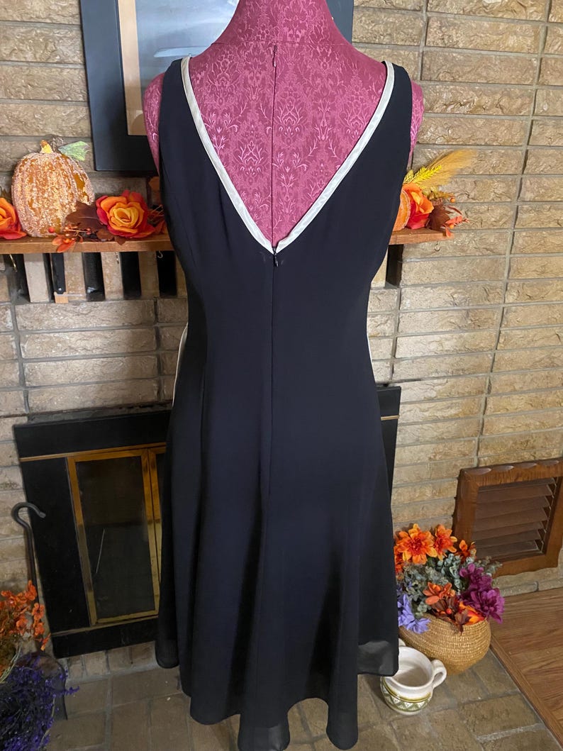 May include: A black sleeveless dress with a V-neckline and white trim. The dress is displayed on a pink dress form. The dress has a flared skirt and a zipper down the back. The background includes a fireplace and fall decorations.