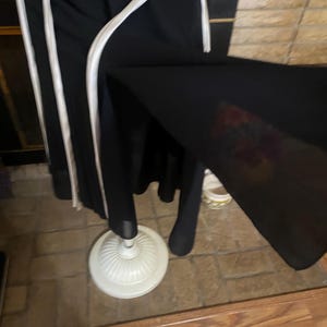 May include: Black garment with white trim hanging on a white dress form. The dress form has a decorative base. The garment appears to be a dress or coat. The background includes a brick wall and wooden floor.