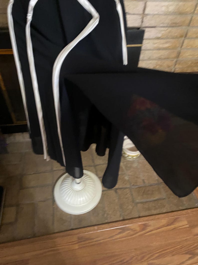 May include: Black garment with white trim hanging on a white dress form. The dress form has a decorative base. The garment appears to be a dress or coat. The background includes a brick wall and wooden floor.