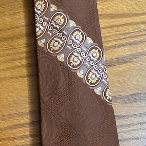 May include: A brown necktie featuring a tonal jacquard pattern of swirling shapes across the fabric. A diagonal band and a bottom border display a repeating decorative motif in white, gold, and brown, consisting of stylized floral elements.