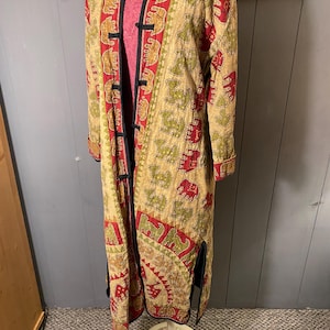 May include: A long, beige, open-front coat featuring a repeating pattern of red and green elephants. The garment includes a mandarin collar, black trim along the front opening, long sleeves with cuffs, and side slits at the hem, displayed on a stand.