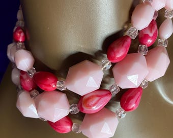 Peut inclure: Collier à plusieurs rangs avec des perles roses et rouges. Le collier présente des perles géométriques facettées et en forme de goutte. De petites perles transparentes séparent les plus grosses. Le collier est présenté sur un mannequin.
