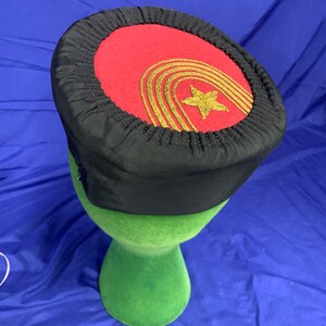 May include: A black and red hat with a gold star and a gold stripe design. The hat is on a green display stand.