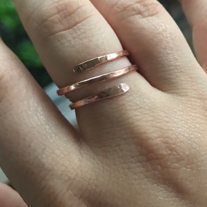 May include: A copper-colored wrap ring with a hammered texture. The ring features a spiral design that wraps around the finger. The metal has a warm, reddish-brown hue, and the surface reflects light.