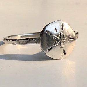 May include: A silver ring featuring a sand dollar design. The ring has a textured band and a detailed sand dollar centerpiece. The ring is a piece of jewelry, perfect for those who love the beach or ocean-themed accessories.