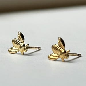 May include: A pair of gold-colored bee stud earrings. Each earring features a detailed bee design with wings and body segments. The earrings are small and delicate, suitable for everyday wear.