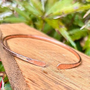 May include: A hammered copper cuff bracelet with an open design. The bracelet is displayed on a wooden surface, with green foliage in the background. The cuff is a simple, elegant piece of jewelry.