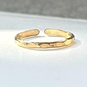 May include: A gold-colored open ring featuring a textured, hammered surface finish. The band is thin and circular with a small gap at the top, creating an adjustable design. The metal surface reflects light, highlighting the uneven, hand-crafted appearance of the hammered texture across the entire outer circumference of the ring.