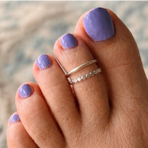 May include: Two silver toe rings worn on the second toe. The top ring is a smooth, plain band with a rounded profile. The bottom ring features a textured, geometric pattern consisting of small, repeating hexagonal shapes around the entire band.