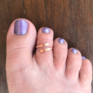 May include: A rose gold toe ring with a simple, minimalist design. The ring is made of two thin bands of metal that are joined together at the top. The ring is worn on the second toe of a foot with purple nail polish.