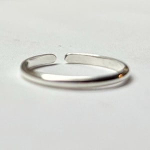 May include: A silver-colored adjustable toe ring. The ring is open at the top, allowing for size adjustment. The ring is smooth and shiny, reflecting light. The background is a plain white surface.