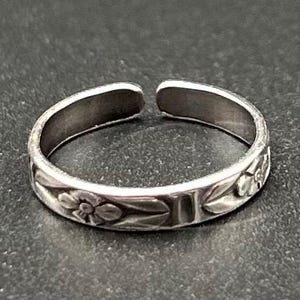 May include: A silver-toned adjustable toe ring with a floral design. The ring features embossed flowers and leaves around the band. The open design allows for size adjustment. The ring is displayed on a dark surface.