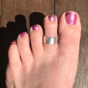 May include: A silver-colored toe ring with a hammered texture is worn on a foot with pink nail polish. The ring is a simple band design, adding a touch of understated elegance to the foot.