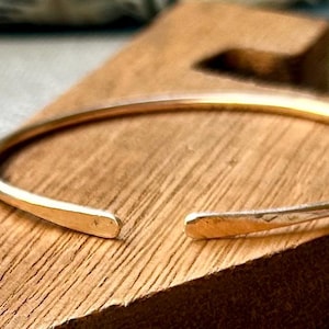 May include: A gold-colored open cuff bracelet resting on a wooden surface. The bracelet has a simple, minimalist design with slightly flattened ends. The wood has a natural grain and a warm, brown tone.