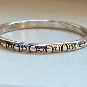 May include: A silver ring with a repeating pattern of small circles and vertical lines. The ring is thin and has a slightly textured surface. The design is consistent around the entire band, creating a simple yet elegant look. This ring is a piece of jewelry.