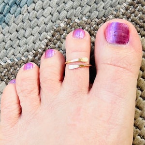 May include: A close-up of a foot with a gold and silver toe ring. The ring wraps around the toe. The toenails are painted with a purple and pink gradient. The background is a gray and brown woven rug.