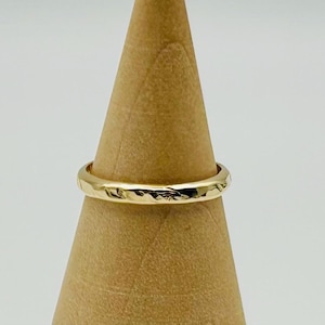 May include: A gold ring with a textured surface is displayed on a wooden cone. The ring has a hammered appearance, reflecting light. The ring is a simple band, likely for everyday wear. The ring is made of gold.