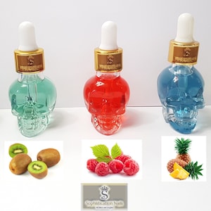 May include: Five small glass bottles shaped like skulls filled with colorful liquids. The bottles have gold lids with droppers. The bottles are arranged in a row from left to right: blue, green, red, blue, and yellow. Below the bottles are images of vanilla beans, kiwi fruit, raspberries, pineapple, and peaches.