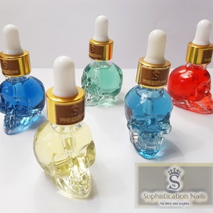 May include: Five glass bottles shaped like skulls filled with colorful liquids. The bottles have gold lids with droppers and are labeled with a gold "S" logo. The bottles are arranged on a white surface. The text on the label reads "Sophistication Nails Au del&agrave; des ongles".