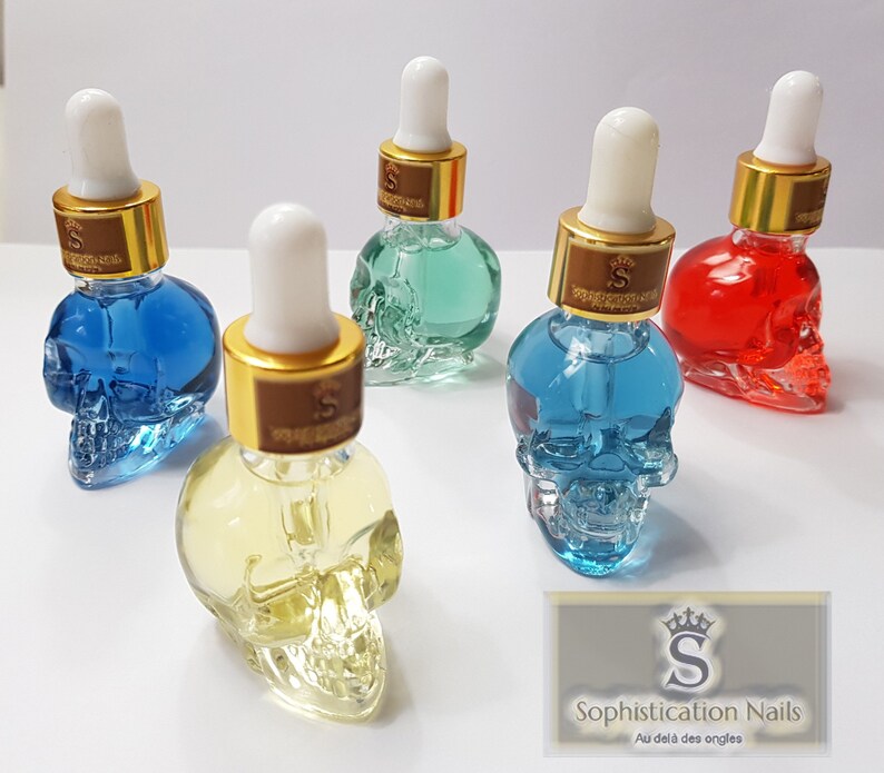 May include: Five glass bottles shaped like skulls filled with colorful liquids. The bottles have gold lids with droppers and are labeled with a gold "S" logo. The bottles are arranged on a white surface. The text on the label reads "Sophistication Nails Au del&agrave; des ongles".