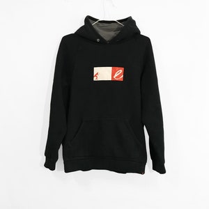 Puede incluir: Sudadera con capucha negra con un logotipo de canguro y la letra "E" en la parte delantera. El logotipo está sobre un fondo rojo, blanco y beige.