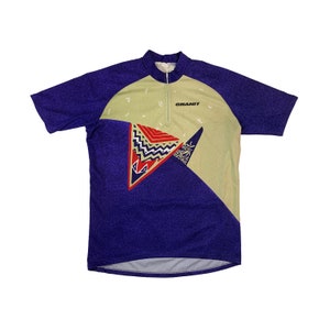 May include: A short-sleeved cycling jersey in a purple and tan colour block design. The jersey has a half-zip closure and the word "GRANIT" printed on the chest. The design includes geometric shapes in red, blue, and white.