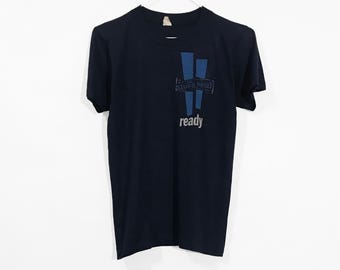 May include: A dark navy blue short-sleeve t-shirt featuring a graphic on the upper right chest. The design includes two vertical blue rectangles, a rectangular box containing the text "Blues Band", and the word "ready" printed in white below.
