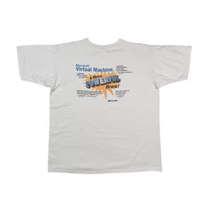 Pode incluir: Camiseta branca com um gráfico azul e amarelo que diz "Microsoft Virtual Machine A More Powerful Brew!" O gráfico apresenta uma explosão no estilo de quadrinhos com as palavras "Powerful Brew!" em uma estrela amarela. O texto abaixo do gráfico inclui informações sobre o produto e o logotipo da Microsoft.