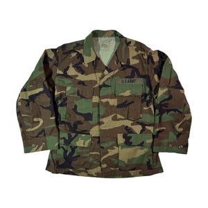 Puede incluir: Chaqueta militar con estampado de camuflaje con el texto "U.S. ARMY" en el bolsillo del pecho. La chaqueta tiene cierre de botones, múltiples bolsillos y un cuello verde. El diseño de camuflaje incorpora tonos de verde, marrón y negro.
