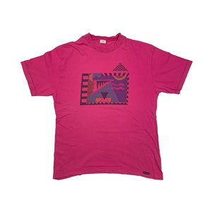 May include: A pink t-shirt with a geometric design in shades of purple, blue, orange, and brown. The design is framed by a black border.