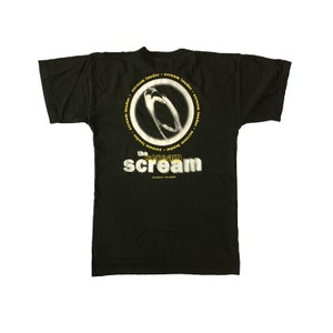 Op de afbeelding: Zwart T-shirt met een witte en gele afbeelding van een schreeuwend gezicht in een cirkel met de tekst "scream louder" die rond de cirkel wordt herhaald. Onder de cirkel staat de tekst "the scream scream".