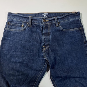 Puede incluir: Primer plano de unos vaqueros de mezclilla azules Carhartt. Los vaqueros tienen bragueta con botones, dise&ntilde;o de cinco bolsillos y un lavado descolorido. El logotipo de Carhartt es visible en la cintura. Los vaqueros son un estilo cl&aacute;sico y duradero.