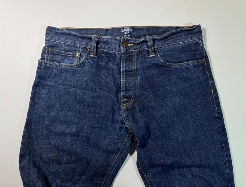 Puede incluir: Primer plano de unos vaqueros de mezclilla azules Carhartt. Los vaqueros tienen bragueta con botones, dise&ntilde;o de cinco bolsillos y un lavado descolorido. El logotipo de Carhartt es visible en la cintura. Los vaqueros son un estilo cl&aacute;sico y duradero.