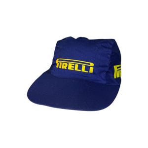 May include: A navy blue cap with a curved brim. The cap features the word "PIRELLI" in yellow block letters across the front. A yellow logo is on the right side of the cap. The cap is made of a durable fabric.