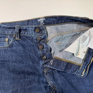 Puede incluir: Primer plano de unos vaqueros Carhartt WIP de color azul. Los vaqueros tienen bragueta con botones, cinco bolsillos y una etiqueta que dice "34/34". El denim es de lavado medio con textura visible.