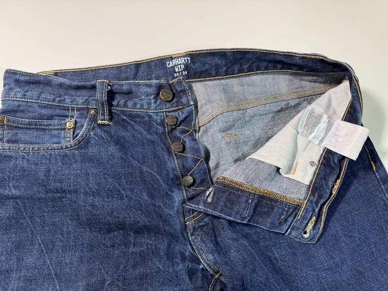 Puede incluir: Primer plano de unos vaqueros Carhartt WIP de color azul. Los vaqueros tienen bragueta con botones, cinco bolsillos y una etiqueta que dice "34/34". El denim es de lavado medio con textura visible.