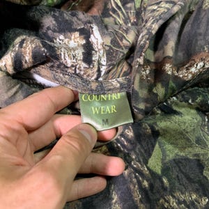 May include: A green, brown and black camouflage print shirt with a green tag that says "COUNTRY WEAR" and "M" in white text.