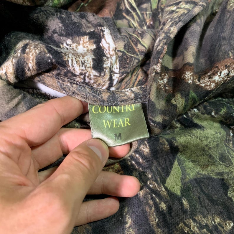 May include: A green, brown and black camouflage print shirt with a green tag that says "COUNTRY WEAR" and "M" in white text.