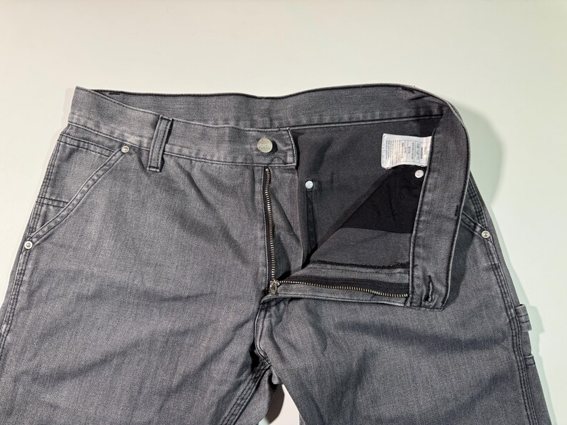 Peut inclure: Pantalon de travail en jean gris avec fermeture &eacute;clair, bouton et poches avant. Le pantalon a une conception classique &agrave; cinq poches et est fait d'un mat&eacute;riau en denim durable. Le pantalon est montr&eacute; d&eacute;boutonn&eacute; et d&eacute;zipp&eacute;.