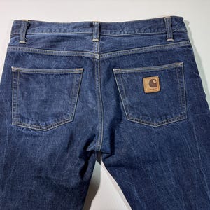 Puede incluir: Vista de cerca de la parte trasera de unos vaqueros azules. Los vaqueros tienen dos bolsillos traseros y un parche de cuero marr&oacute;n con un logotipo en el bolsillo derecho. La mezclilla tiene un aspecto ligeramente descolorido.