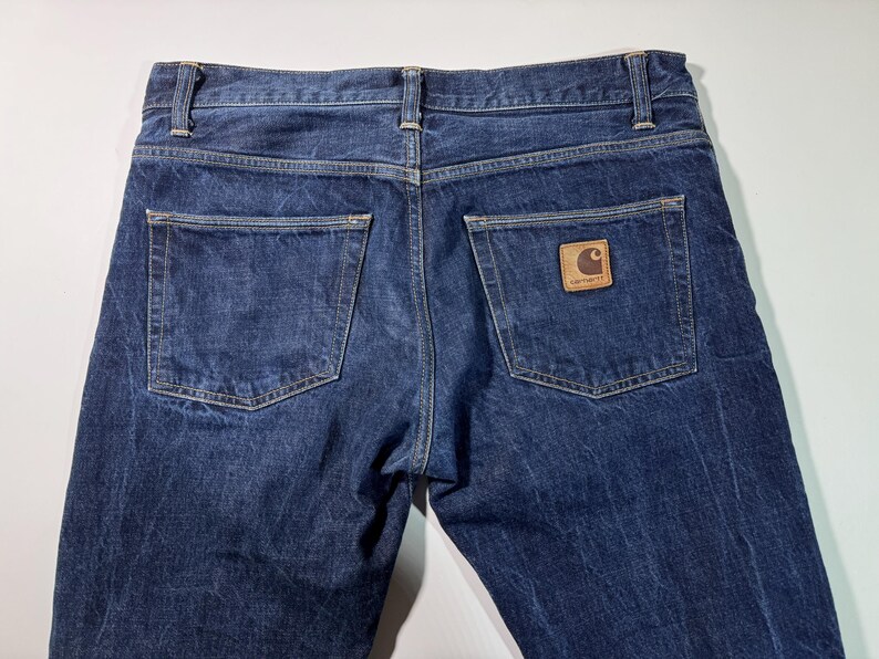 Puede incluir: Vista de cerca de la parte trasera de unos vaqueros azules. Los vaqueros tienen dos bolsillos traseros y un parche de cuero marr&oacute;n con un logotipo en el bolsillo derecho. La mezclilla tiene un aspecto ligeramente descolorido.